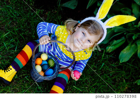 Cute little toddler girl with bunny ears having fun with traditional Easter eggs hunt on warm sunny day, outdoors. Happy child celebrating family christian holiday with basket with colored egg Cute little toddler girl with bunny ears having fun with traditional Easter eggs hunt on warm sunny day, outdoors. Happy child celebrating family christian holiday with basket with colored egg 110278398
