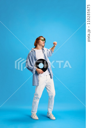 Body size portrait of young man, dressed casual outfit dancing with vintage vinyl record disk against blue studio background. 110285675