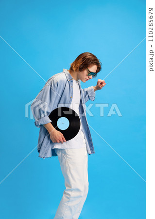 Side view portrait of happy, overjoyed man, student in modern sunglasses dancing with vinyl record against blue studio background. 110285679