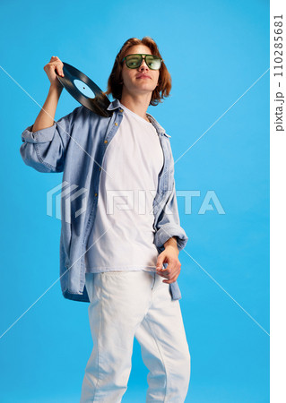 Cool attitude man, student in stylish sunglasses holding vinyl record, disk against blue studio background. Retro music. 110285681