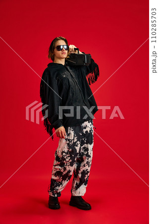 Full-length portrait of young, well-dressed man listening popular music from vintage tape recorder against red studio background. 110285703