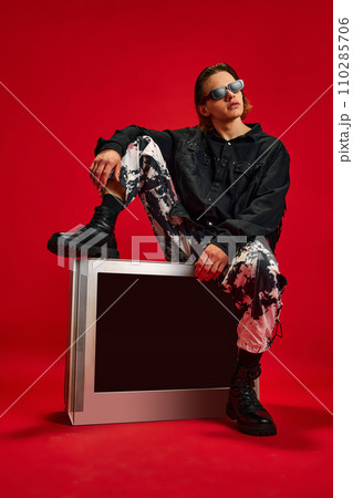 Young man in style clothes, looks as cowboy sitting on retro TV and looking away against red studio background. Concept of people and technology. 110285706