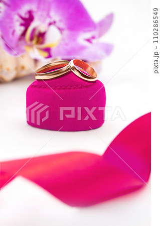 Gold wedding rings on pink velvet heart on gift box with orchids on white background. Marriage, proposal, Valentine's Day. Vertical. Copy space Gold wedding rings on pink velvet heart on gift box with orchids on white background. Marriage, proposal, Valentine's Day. Vertical. Copy space 110286549