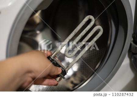 Close-up of a hand holding a heating element covered with limescale. Washing machine failure due to scale accumulation on the heating element. Washing machine heating element repair concept. Close-up of a hand holding a heating element covered with limescale. Washing machine failure due to scale accumulation on the heating element. Washing machine heating element repair concept. 110287432