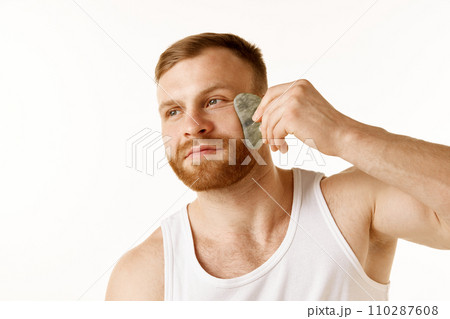 Portrait of young man in T-shirt, taking care of his facial skin using massage scrapper against white background. Concept of anti-aging procedures. 110287608