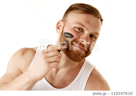 Young man takes care of his facial skin, removes signs of aging and tightens his facial contours using massage roller against white background. 110287610