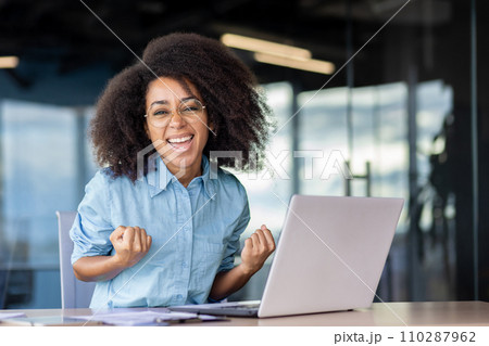 Portrait of a happy and successful young business woman sitting in the office in front of the laptop and looking happy at the camera, celebrating and showing a victory gesture with her hands. 110287962