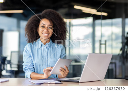 Portrait of a smiling young African American woman sitting at a desk in the office, holding a tablet and smiling at the camera. 110287970