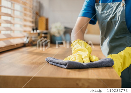 Maid in protective glove wipes wood table in cozy kitchen. Using professional cleaning products for perfect home tidiness. Cleaner working safety glove hygiene routine. Maid housekeeping concept. Maid in protective glove wipes wood table in cozy kitchen. Using professional cleaning products for perfect home tidiness. Cleaner working safety glove hygiene routine. Maid housekeeping concept. 110288778