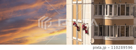 Repair work at height (sealing seams in a panel house), against the sky at sunset. Worker insulates the seams of the facade of an old residential building. Maintenance of residential buildings Repair work at height (sealing seams in a panel house), against the sky at sunset. Worker insulates the seams of the facade of an old residential building. Maintenance of residential buildings 110289596