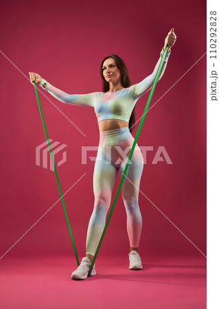 Confident female with dark hair, doing arm exercise with resistance band, while working out. Low angle view of fit woman in sportswear exercising, against pink background. Fitness and sport concept. 110292828