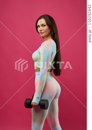 Gorgeous fit woman posing with weights dumbbells, looking at camera with light smile. View from shoulder of confident Caucasian female athlete working out, isolated on pink background. Sport concept. Gorgeous fit woman posing with weights dumbbells, looking at camera with light smile. View from shoulder of confident Caucasian female athlete working out, isolated on pink background. Sport concept. 110292842