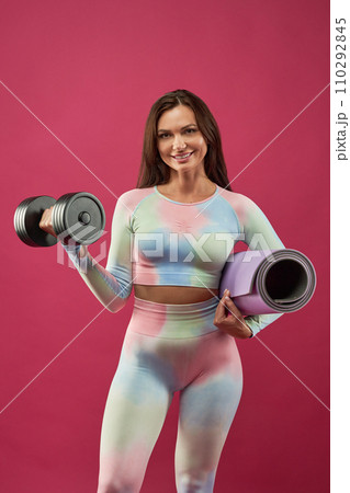 Cheerful Caucasian girl in light activewear, raising dumbbell, while looking at camera. Portrait of nice lady standing with yoga mat, having workout, isolated on pink studio background. Sport concept. Cheerful Caucasian girl in light activewear, raising dumbbell, while looking at camera. Portrait of nice lady standing with yoga mat, having workout, isolated on pink studio background. Sport concept. 110292845