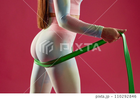 Close up of anonymous sexy female athlete exercising, strengthen buttocks with rubber band indoors. Crop view of seductive woman with green elastic band, working out, isolated on pink. Sport concept. 110292846