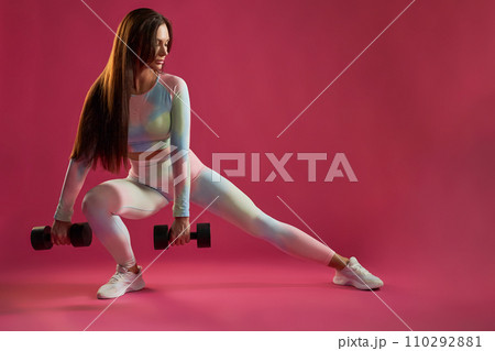 Strong brunette female in leggings stretching leg during fitness workout indoors. Front view of motivated middle-aged woman doing side lunge with dumbbells, against hot pink background. Sport concept. 110292881