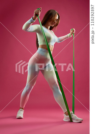 Slim Caucasian sportswoman stepping, pulling resistance rope, during fitness training indoors. Low angle view of focused young girl in sportswear, doing arm exercise, isolated on pink. Fitness concept 110292897