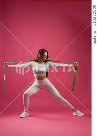 Strong female in sportswear stretching with rubber band during fitness workout indoors. Front view of motivated middle-aged woman doing lung with sport rope, against pink background. Sport concept. Strong female in sportswear stretching with rubber band during fitness workout indoors. Front view of motivated middle-aged woman doing lung with sport rope, against pink background. Sport concept. 110292909