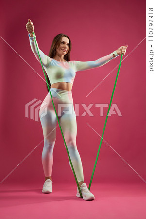 Female fitness trainer doing arm exercise with resistance band, working out indoors. Low angle view of attractive middle aged woman in legging exercising, against pink background. Fitness concept. Female fitness trainer doing arm exercise with resistance band, working out indoors. Low angle view of attractive middle aged woman in legging exercising, against pink background. Fitness concept. 110292918