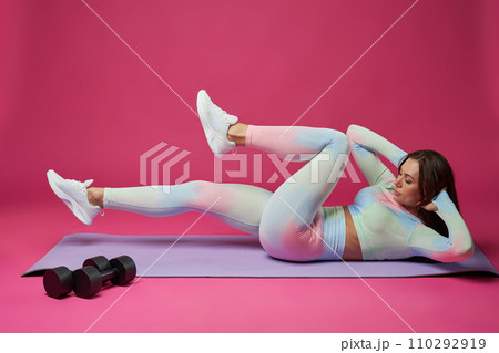 Focused woman in activewear keeping hands behind head, having abdominal crunch workout. Side view of fit female doing exercise for core, lying on math against pink studio background. Sport concept. 110292919