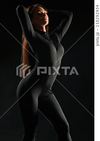 Graceful, relaxed woman wearing black, dancing in low light Illumination. Side view of beauty posing, gently touching long hair, demonstrating body shapes, against black background. Beauty concept. 110292934