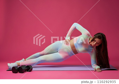 Brunette fit female in light sportswear doing side plank exercise while training in studio. Side view of fit determined female exercising alone on mat, isolated on pink background. Concept of sport. Brunette fit female in light sportswear doing side plank exercise while training in studio. Side view of fit determined female exercising alone on mat, isolated on pink background. Concept of sport. 110292935