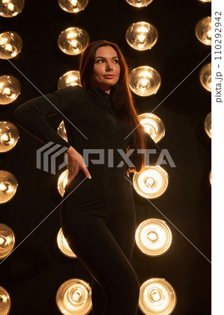 Stunning lady wearing dark apparel, posing with hand on waist in black studio. Front view of Caucasian woman with long hair, pensively looking away, in front of spot lights background. Beauty concept. 110292942