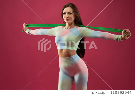 Strong, beautiful woman doing rubber band exercises in studio. Portrait of smiling female athlete with spread arms, exercising with elastic band, looking at camera, isolated on pink. Sport concept. 110292944