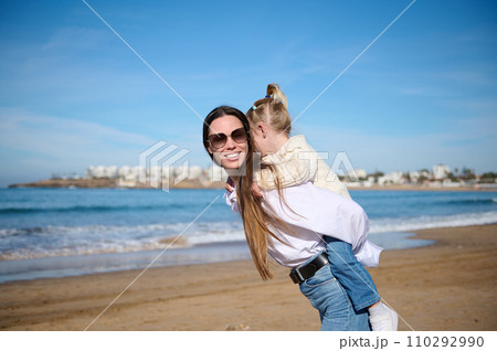Happy family of a young loving caring mother carrying little daughter on her back and walking on tropical sandy beach. Happy family of a young loving caring mother carrying little daughter on her back and walking on tropical sandy beach. 110292990