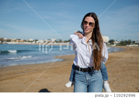 Happy Caucasian young mother carrying her little daughter on her back at the beach under a blue sky in the morning Happy Caucasian young mother carrying her little daughter on her back at the beach under a blue sky in the morning 110292996