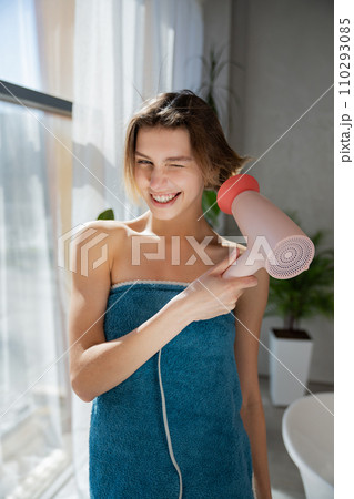 Smiling young woman wearing tower, posing, while drying wet hair beside window. Front view of beautiful girl with haircut winking to camera in bathroom in sunny morning. Lifestyle, routine concept.  110293085
