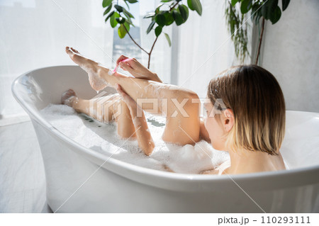 Young caucasian girl with haircut, shaving leg, while lying in bath at home. Back view of slim woman using razor to remove extra hair from body, while bathing in sunlit bathroom. Body care concept. 110293111