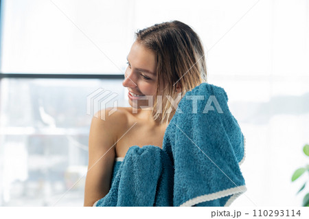 Happy young female drying hair with turquoise cotton towel after showering against wide window. Portrait of smiling caucasian woman using towel on wet head in bathroom. Concept of hair care, beauty. 110293114