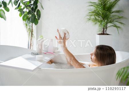 Relaxed caucasian lady washing arms with foamy sponge, while bathing. Side view of young caucasian woman lather body with washcloth, while soaking in bathtub, surrounded by houseplant. Concept of spa. Relaxed caucasian lady washing arms with foamy sponge, while bathing. Side view of young caucasian woman lather body with washcloth, while soaking in bathtub, surrounded by houseplant. Concept of spa. 110293117