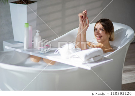 Smiling beautiful girl washing hands, while resting in bathtub. Side view of happy female with haircut taking bath, with set of bath accessories on bath table, with sunbeams on face. Hygiene concept. Smiling beautiful girl washing hands, while resting in bathtub. Side view of happy female with haircut taking bath, with set of bath accessories on bath table, with sunbeams on face. Hygiene concept. 110293126