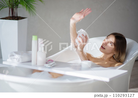 Pretty caucasian woman playing with sun beam, while bathing at home. Side view of joyful blonde girl washing arms, while laying in comfortable bathtub, in front of grey wall. Body care concept.  110293127