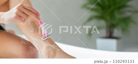 Unrecognizable woman shaving leg with pink razor at home. Close up of anonymous girl removing extra hair, while resting one leg on bathtub edge in bathroom, with blurred background. Body care concept. Unrecognizable woman shaving leg with pink razor at home. Close up of anonymous girl removing extra hair, while resting one leg on bathtub edge in bathroom, with blurred background. Body care concept. 110293131