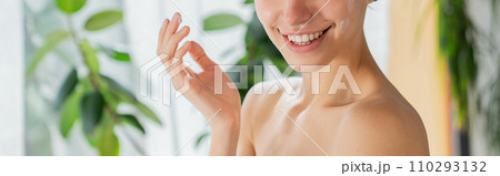 Unrecognizable smiling woman applying face cream after taking bath. Crop view of happy female moisturizing face skin after shower, against blurred houseplants background. Skin and beauty care concept. Unrecognizable smiling woman applying face cream after taking bath. Crop view of happy female moisturizing face skin after shower, against blurred houseplants background. Skin and beauty care concept. 110293132