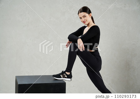 Pretty sporty woman stepping on black cube for fitness and smiling. Side view of fit dark-haired girl with ponytail posing with crossed hands while exercising in studio. Concept of sporty lifestyle. 110293173