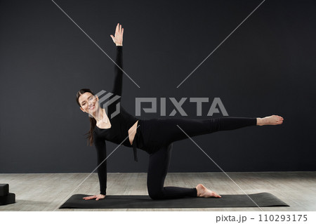 Slender brunette girl practicing pose of yoga, stretching on floor, in front of dark wall background. Front view of attractive woman in activewear exercising alone, looking camera. Concept of yoga. 110293175
