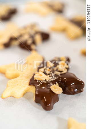 Creating cutout sugar cookies, partially dipped in chocolate and topped with hazelnut pieces, placed on parchment paper. Creating cutout sugar cookies, partially dipped in chocolate and topped with hazelnut pieces, placed on parchment paper. 110294517