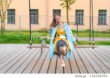 Happy woman swinging on swing in city park, wearing yellow dress and blue coat 110298765