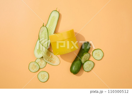 Top view of an unlabeled yellow cosmetic jar and thin cucumber slices on a pastel background. Minimalist cosmetic mockup for advertising. Empty label for branding. 110300512