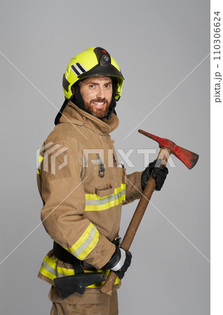 Smiling male firefighter in full gear holding big axe for work. Side view of bearded fireman posing with wooden hatchet tool, while looking at camera, on gray background. Job, equipment, tool concept. Smiling male firefighter in full gear holding big axe for work. Side view of bearded fireman posing with wooden hatchet tool, while looking at camera, on gray background. Job, equipment, tool concept. 110306624