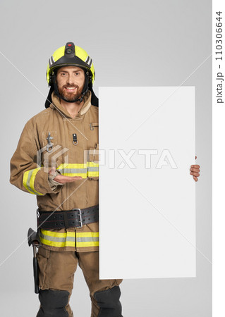 Happy firefighter in uniform standing with blank cardboard indoors. Front view of smiling caucasian fireman demonstrating empty banner, isolated on gray background. Concept of announcement. 110306644