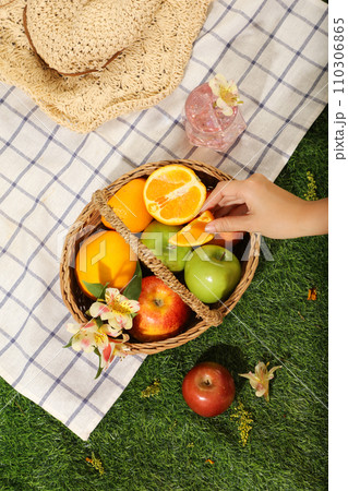 Top view of bamboo basket full with fruits: oranges, apple and green apples. A hand is holding an orange slices. Hat, cool of water cup and checkered decorated for picnic day Top view of bamboo basket full with fruits: oranges, apple and green apples. A hand is holding an orange slices. Hat, cool of water cup and checkered decorated for picnic day 110306865