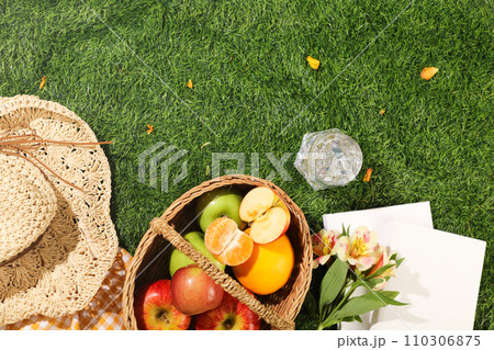 Delicious picnic spread with basket of fruits, hat, cup of cool water and white notebooks on green grass background. Top view, holiday summer concept. Space for design Delicious picnic spread with basket of fruits, hat, cup of cool water and white notebooks on green grass background. Top view, holiday summer concept. Space for design 110306875