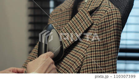 Woman steams plaid jacket with electric steamer at home. Dry cleaning clothes. Household and home equipment for housework. 110307399