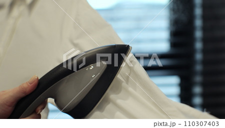 Unrecognizable women hand using handheld steamer on women white shirt, smoothing out creases at home. 110307403