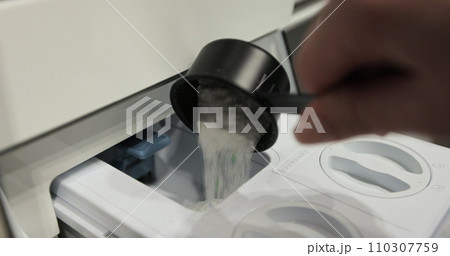 Woman pours washing powder into the washing machine compartment. Female with measuring spoon, fills the washing machine with powder. Woman pours washing powder into the washing machine compartment. Female with measuring spoon, fills the washing machine with powder. 110307759