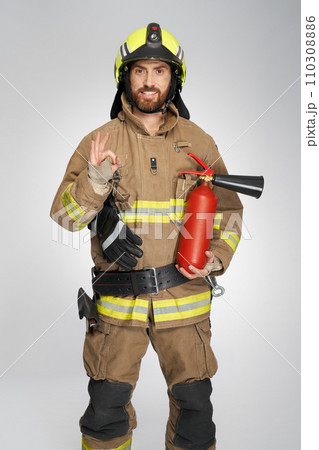 Happy firefighter with extinguisher showing okay sign indoors. Front view of bearded fireman in uniform smiling, while gesturing ok, on gray studio background. Firefighting, hand gesture concept. Happy firefighter with extinguisher showing okay sign indoors. Front view of bearded fireman in uniform smiling, while gesturing ok, on gray studio background. Firefighting, hand gesture concept. 110308886